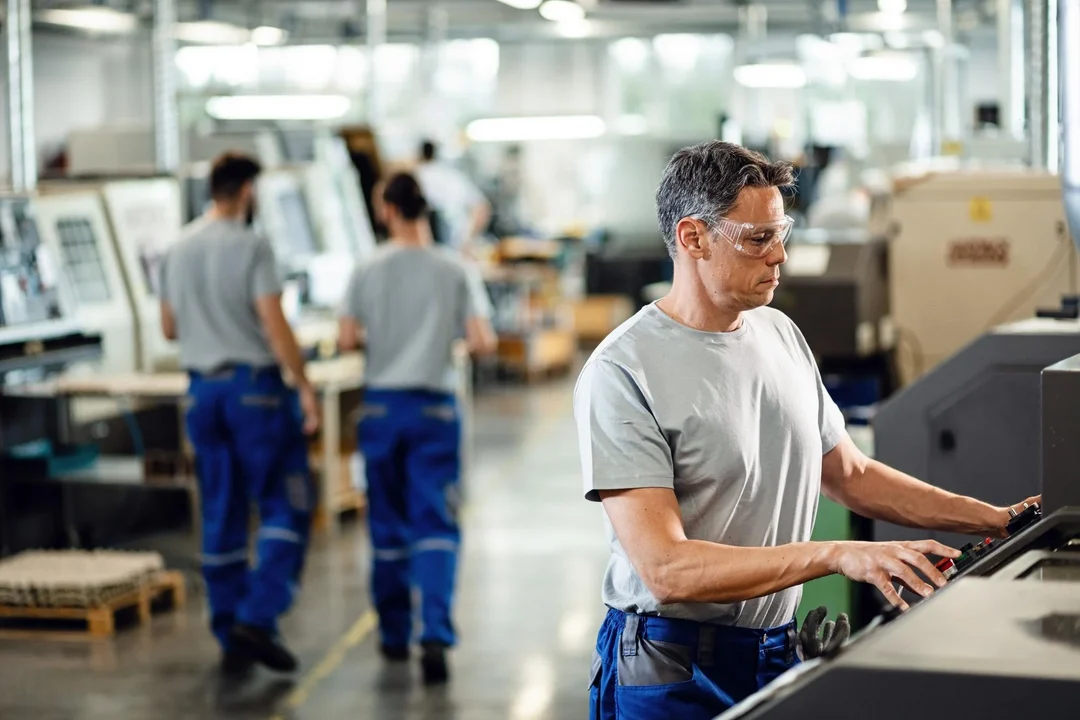 Machinist in safety glasses at a machine control panel, other operators working further down the shop